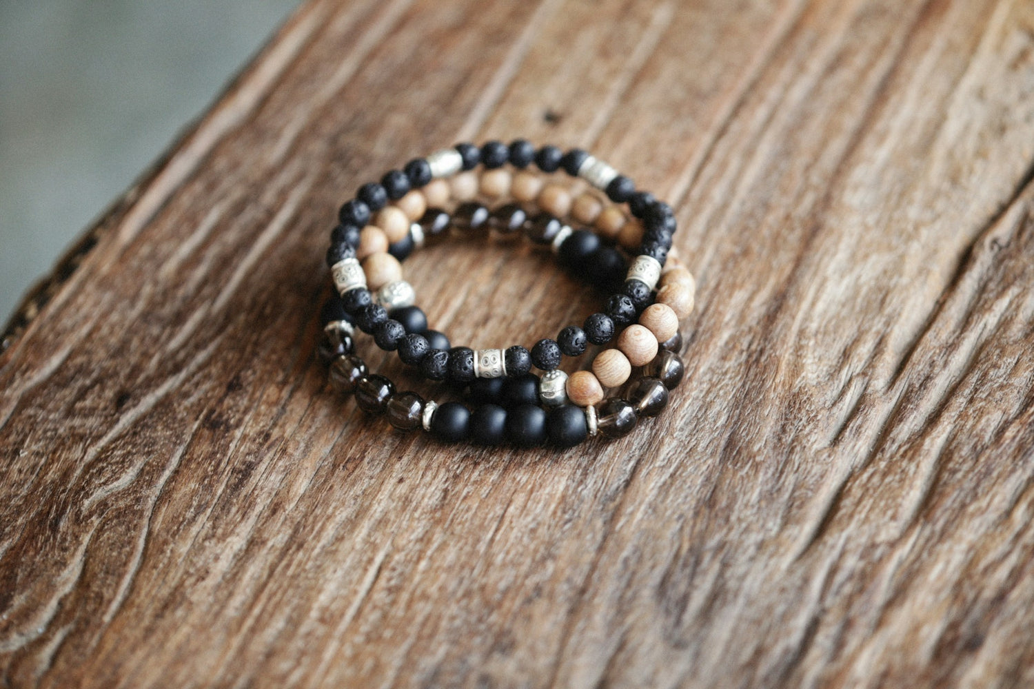 Auf einem Holztisch liegen mehrere Edelstein Armbänder in Naturfarben, gefertigt aus schwarzen Lavasteinen, Holzperlen und silbernen Elementen, die eine schlichte Eleganz ausstrahlen.