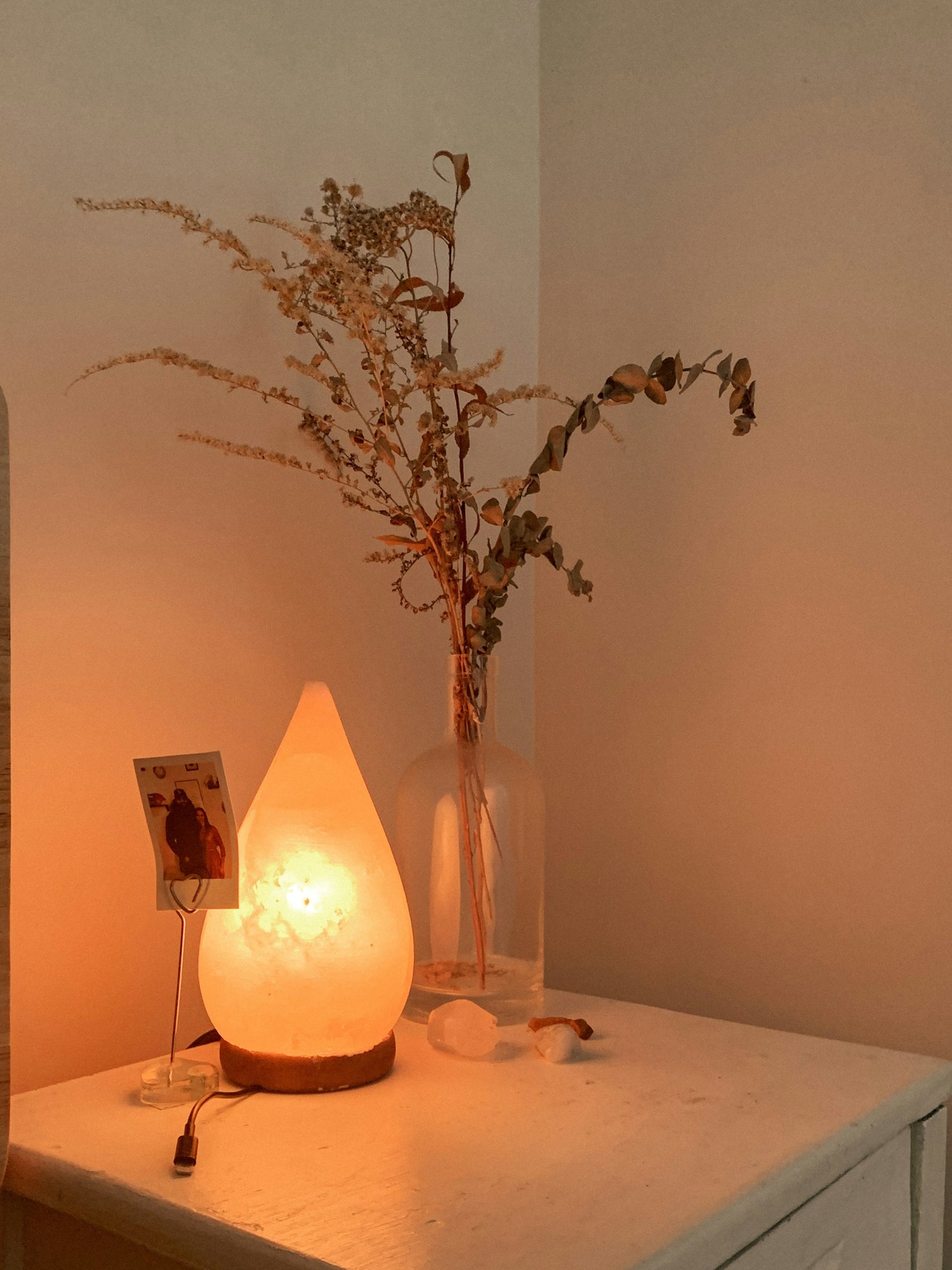 Salzlampe in Tropfenform auf einem weißen Nachttisch, im Hintergrund eine Vase mit getrockneten Zweigen und warmes, sanftes Licht.