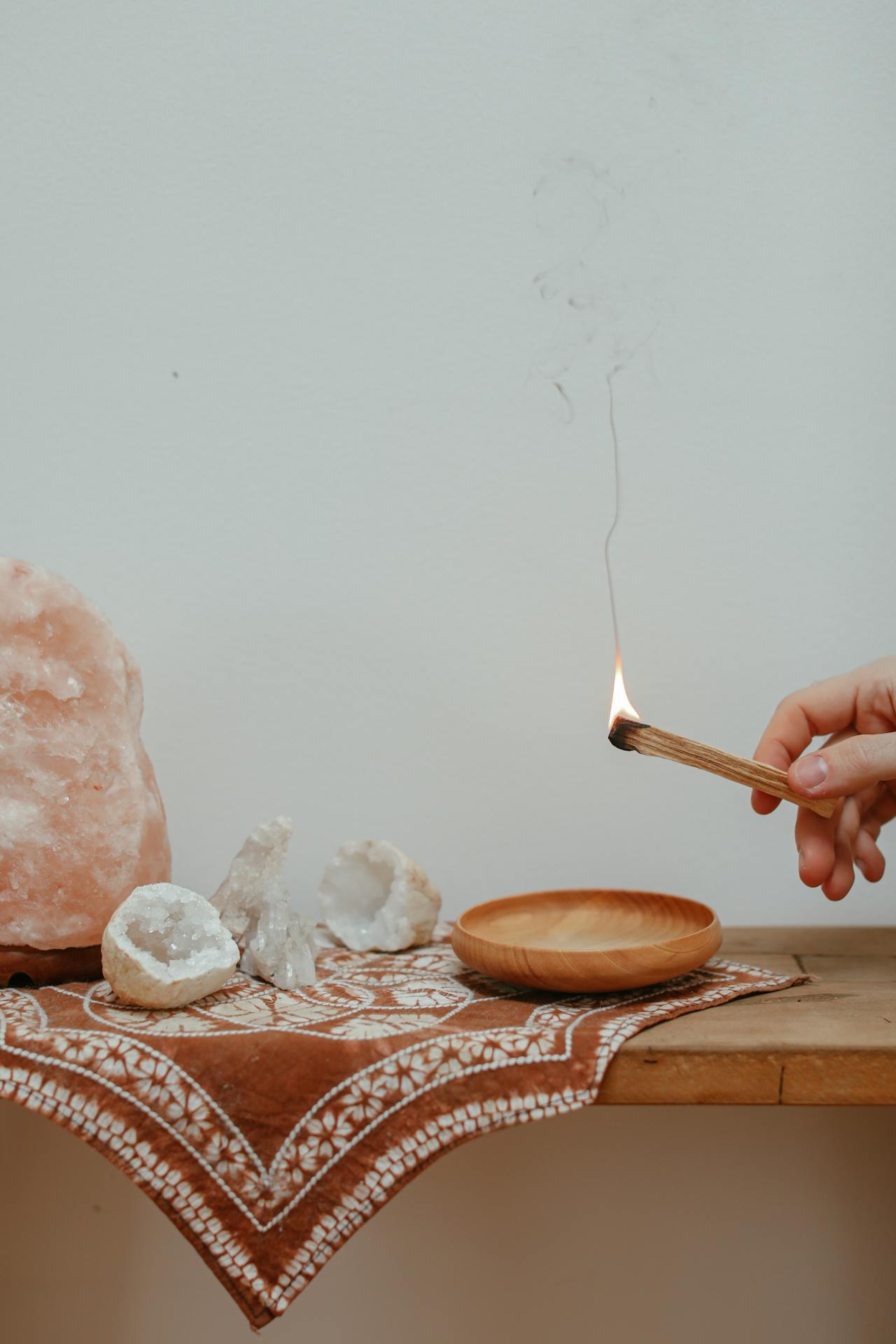 Eine warme Salzlampe leuchtet im Hintergrund auf einem Holztisch, daneben brennt ein Palo-Santo-Stäbchen und liegen funkelnde Kristall-Geoden.