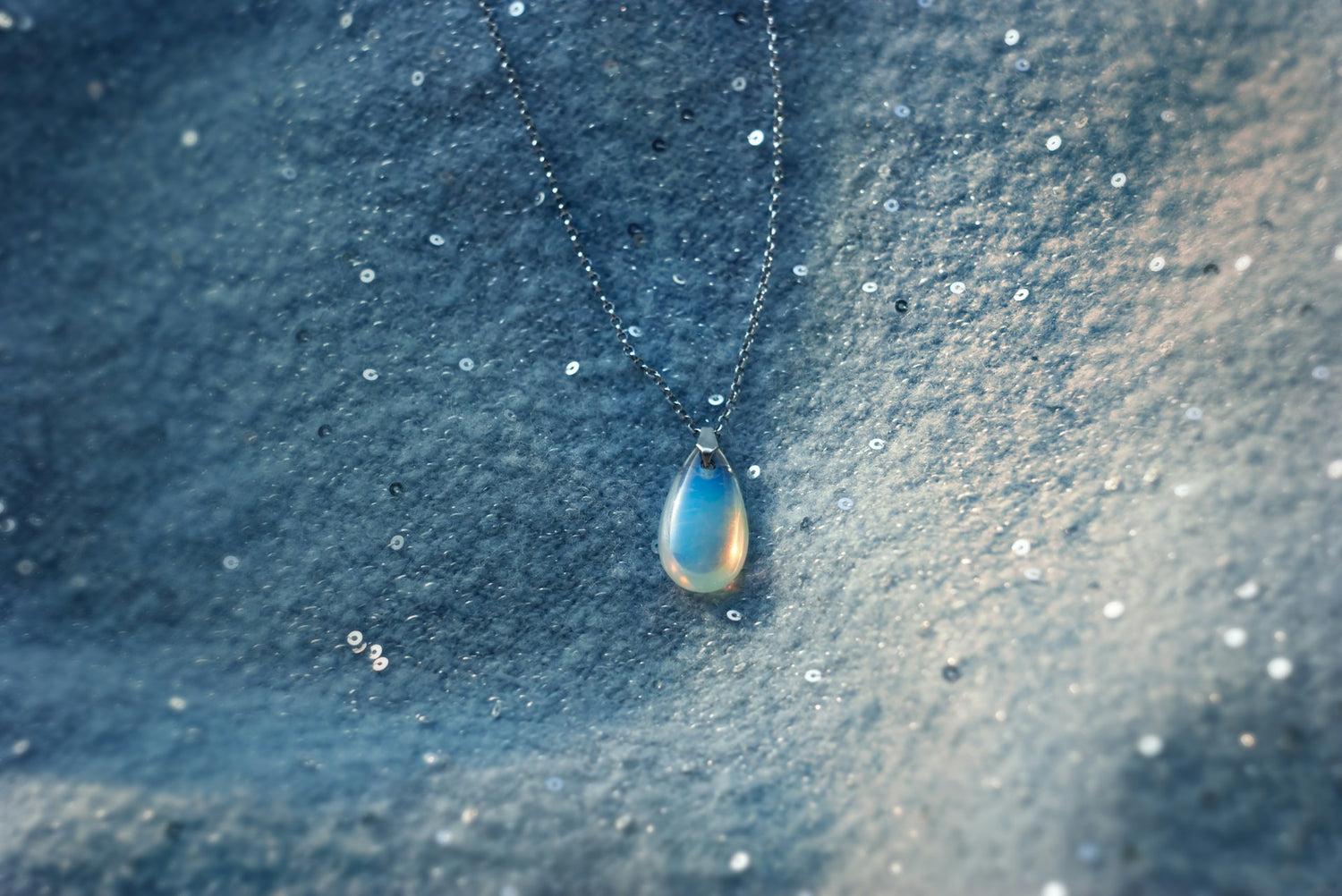 Schimmernder Edelstein Anhänger aus Opal an feiner Silberkette auf funkelndem, blauem Stoff.
