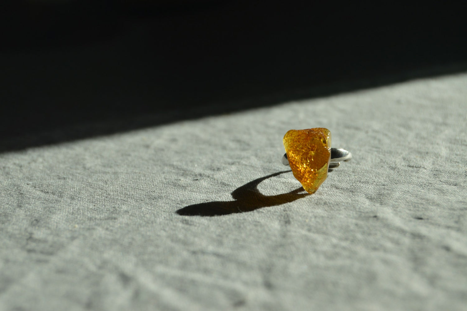 Ein silberner Ring mit einem ungeschliffenen Bernstein Schmuckstein liegt auf einem Stoff, während das Licht seinen warmen Glanz betont.