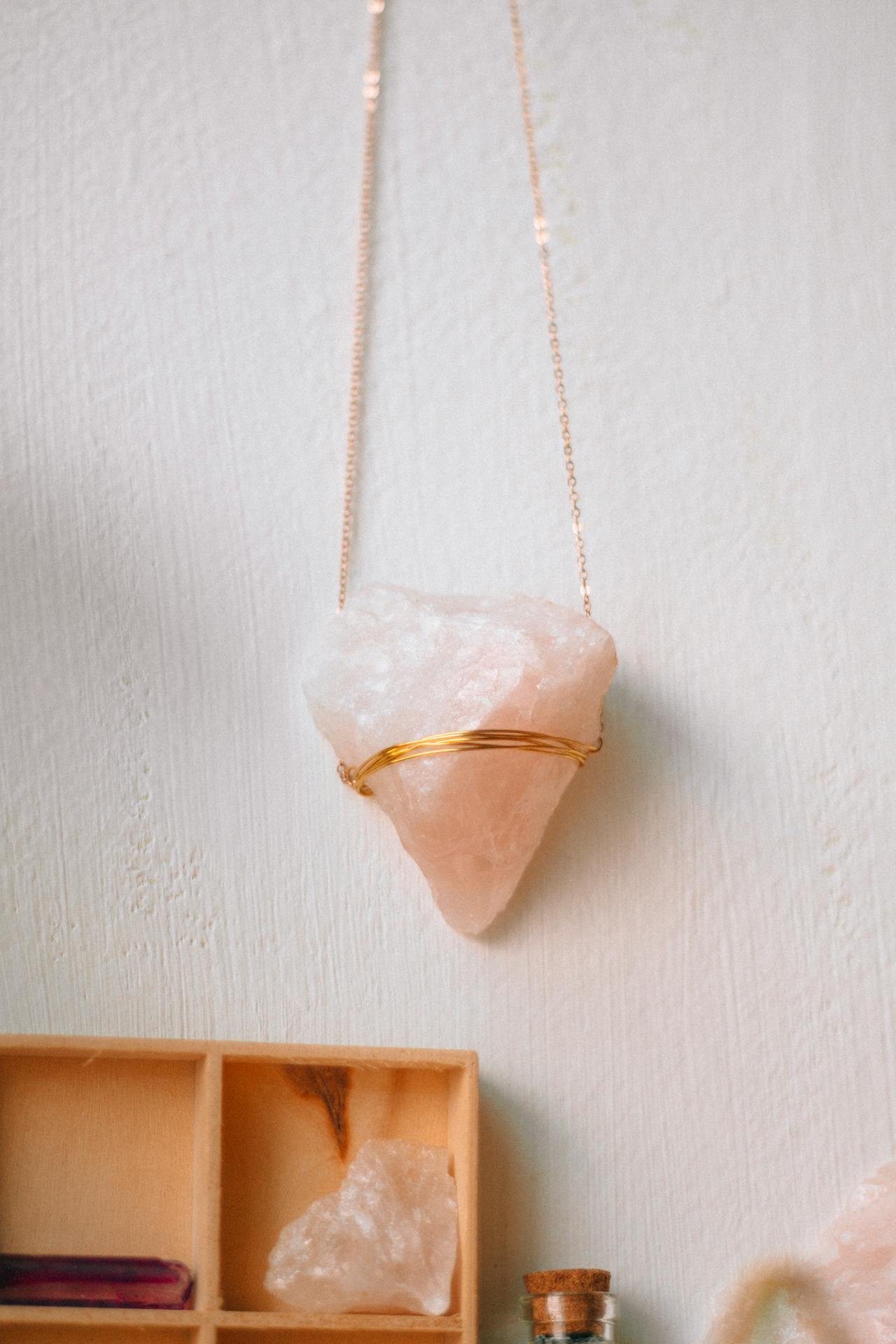 Ein einzigartiger Rosenquarz Schmuck-Anhänger mit goldener Fassung hängt an einer feinen Kette vor einer weißen Wand.
