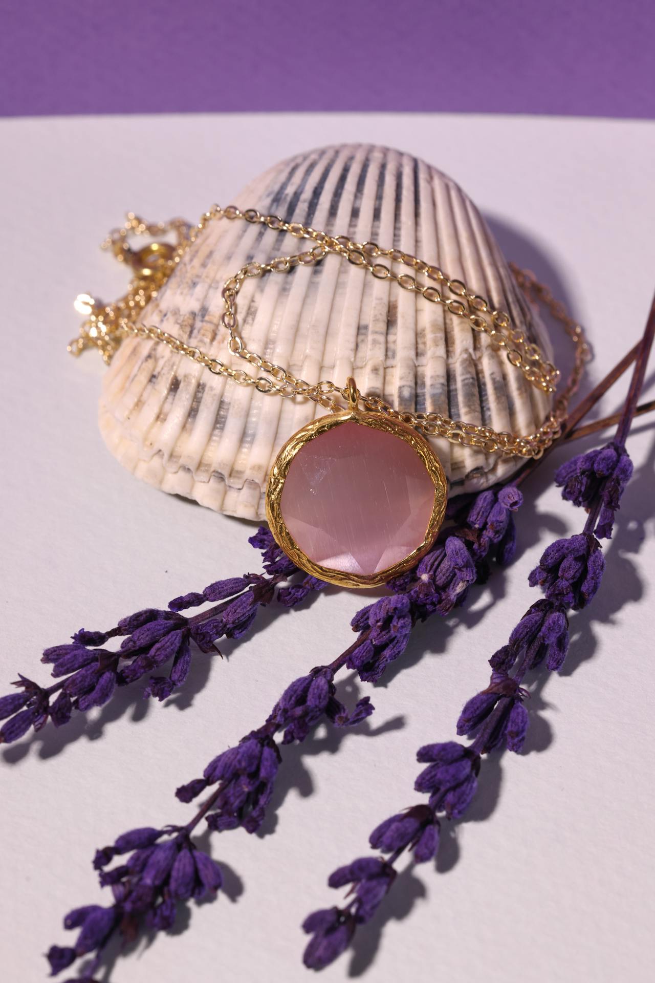 Ein eleganter Rosenquarz Schmuck-Anhänger mit goldener Kette liegt dekorativ auf einer Muschel, umgeben von Lavendelzweigen.