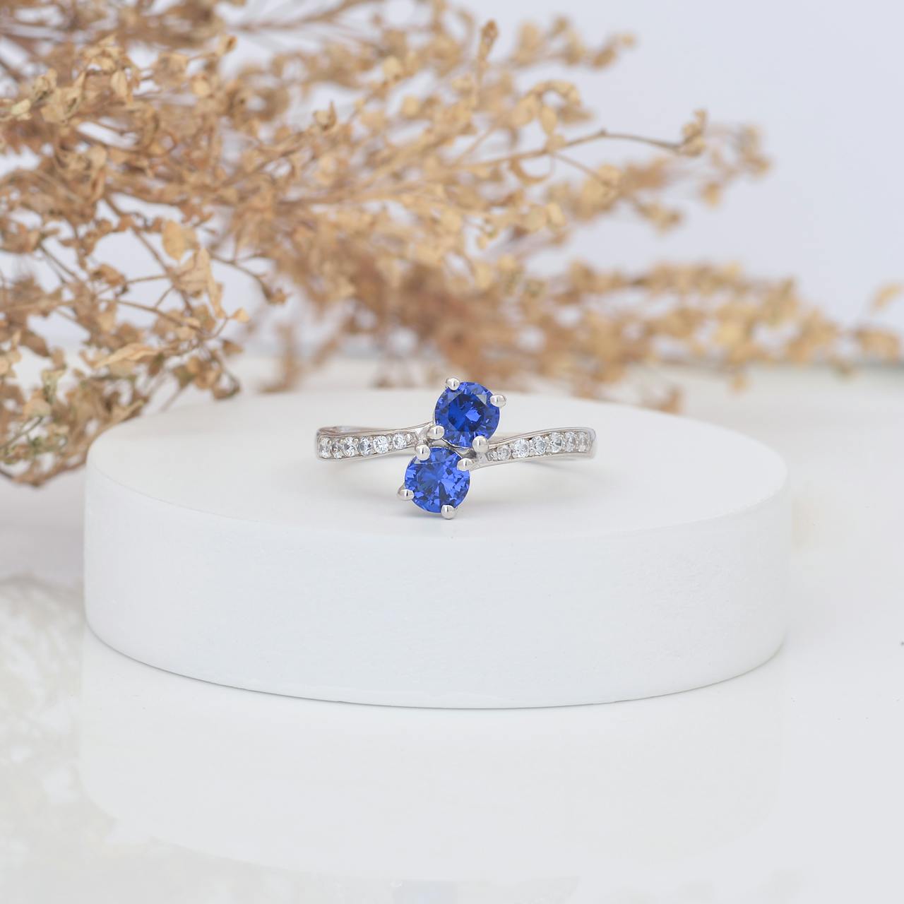 Eleganter Silberring mit funkelndem Saphir Schmuck, stilvoll präsentiert auf einer weißen Plattform mit Trockenblumen im Hintergrund.